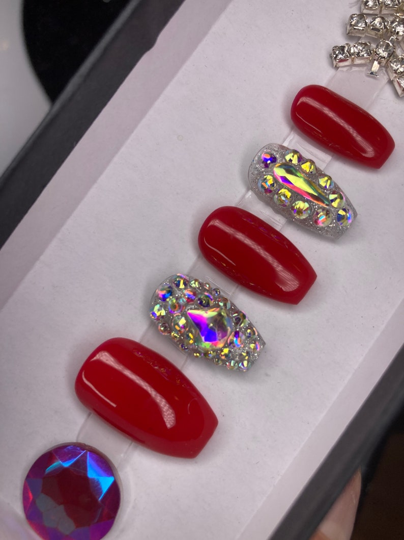 May include: A set of red press-on nails with two accent nails. The accent nails are covered in iridescent rhinestones. A large, faceted, iridescent gem is at the bottom left. Additional rhinestones are at the top right.