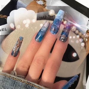 May include: Close-up of a hand with long, blue and white marble-patterned acrylic nails with gold and silver accents. The nails are decorated with rhinestones and small chains.