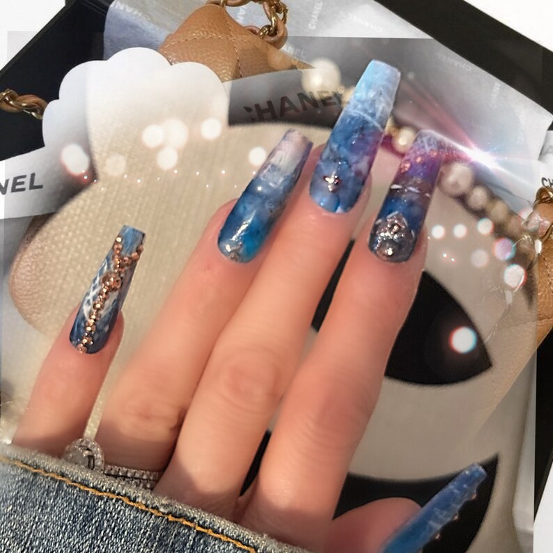 May include: Close-up of a hand with long, blue and white marble-patterned acrylic nails with gold and silver accents. The nails are decorated with rhinestones and small chains.