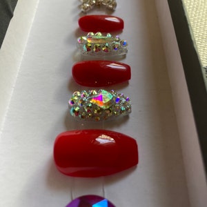 May include: A set of red press-on nails with various rhinestone embellishments. The set includes solid red nails and nails adorned with clear and iridescent rhinestones, arranged in a box.