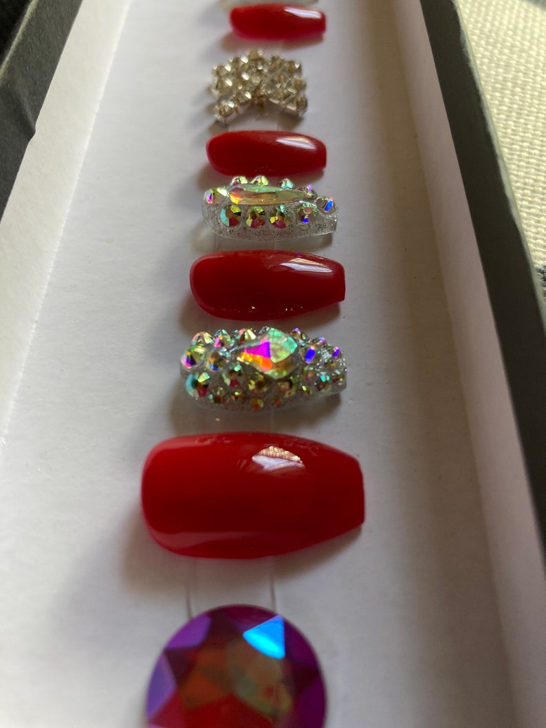 May include: A set of red press-on nails with various rhinestone embellishments. The set includes solid red nails and nails adorned with clear and iridescent rhinestones, arranged in a box.