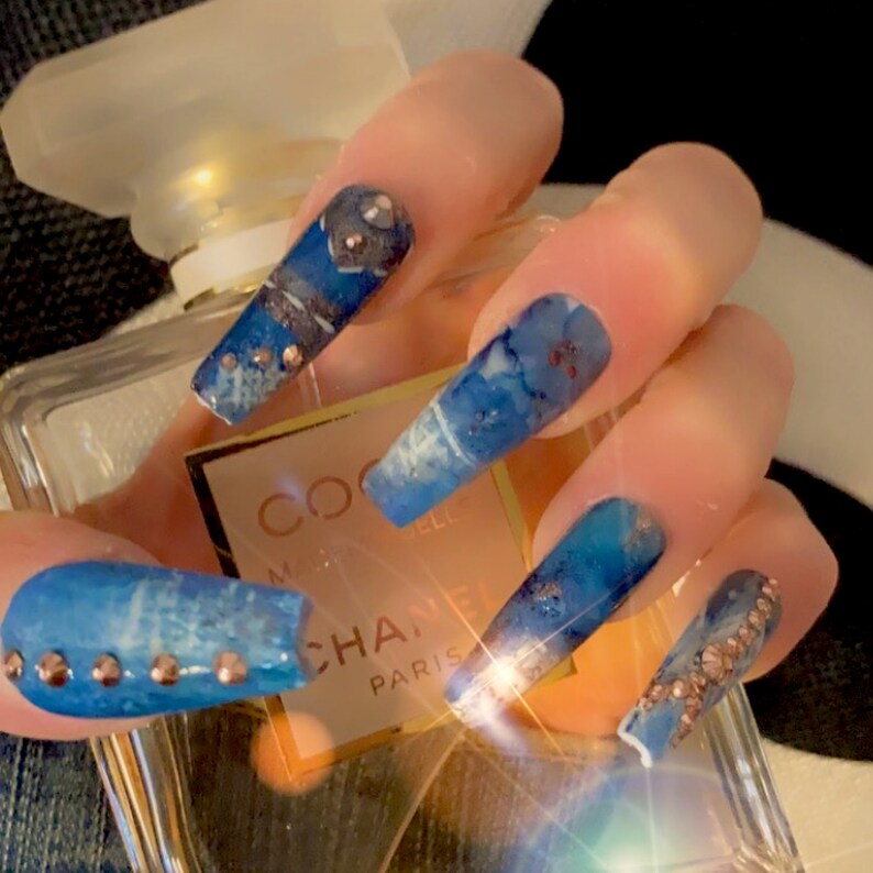 May include: Blue and white marble-effect press-on nails with gold glitter and rhinestones. The nails are long and coffin-shaped. The nails are displayed on top of a bottle of perfume.