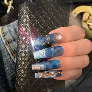 May include: Long, blue and white nail tips with a marble effect and gold accents. The nails are decorated with rhinestones.