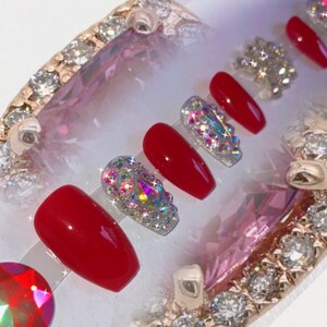 May include: A set of red and bejeweled press-on nails. The nails are a vibrant red color, with some adorned with sparkling, iridescent rhinestones. The nails are arranged on a clear display, with a decorative, jeweled border.