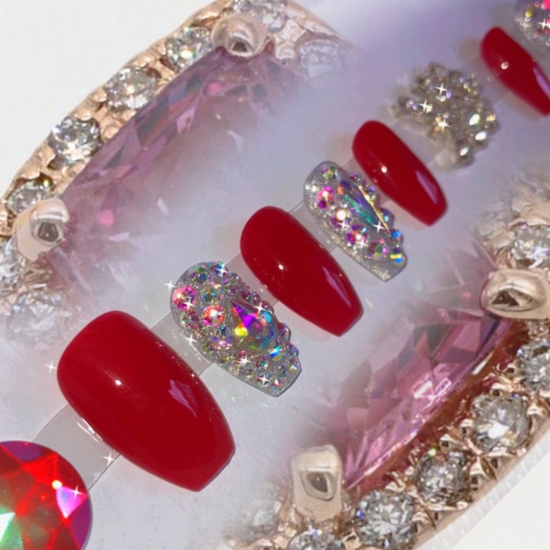 May include: A set of red and bejeweled press-on nails. The nails are a vibrant red color, with some adorned with sparkling, iridescent rhinestones. The nails are arranged on a clear display, with a decorative, jeweled border.
