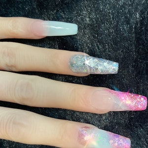 May include: Close-up of long, clear acrylic nails with a gradient of pink and white glitter. The nails are decorated with silver glitter and iridescent glitter.