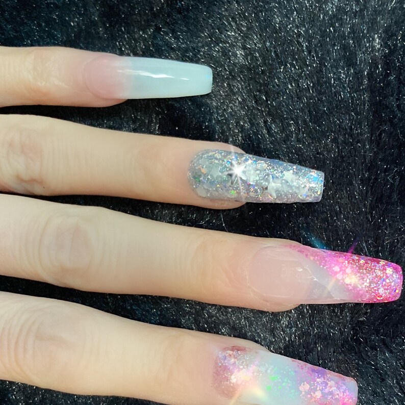 May include: Close-up of long, clear acrylic nails with a gradient of pink and white glitter. The nails are decorated with silver glitter and iridescent glitter.