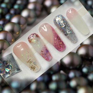 May include: Five long, pink and white ombre press-on nails with glitter accents. One nail features the Chanel logo.