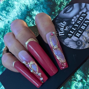 May include: Long, red, coffin-shaped nails with gold accents and iridescent butterfly designs. The nails are decorated with rhinestones and the text "Nails by Allyson Rubin" is visible in the background.