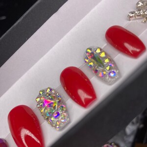 May include: A set of red and rhinestone-encrusted press-on nails. The nails are displayed in a black box with a white interior. The red nails have a glossy finish, while the rhinestone nails sparkle with iridescent gems.