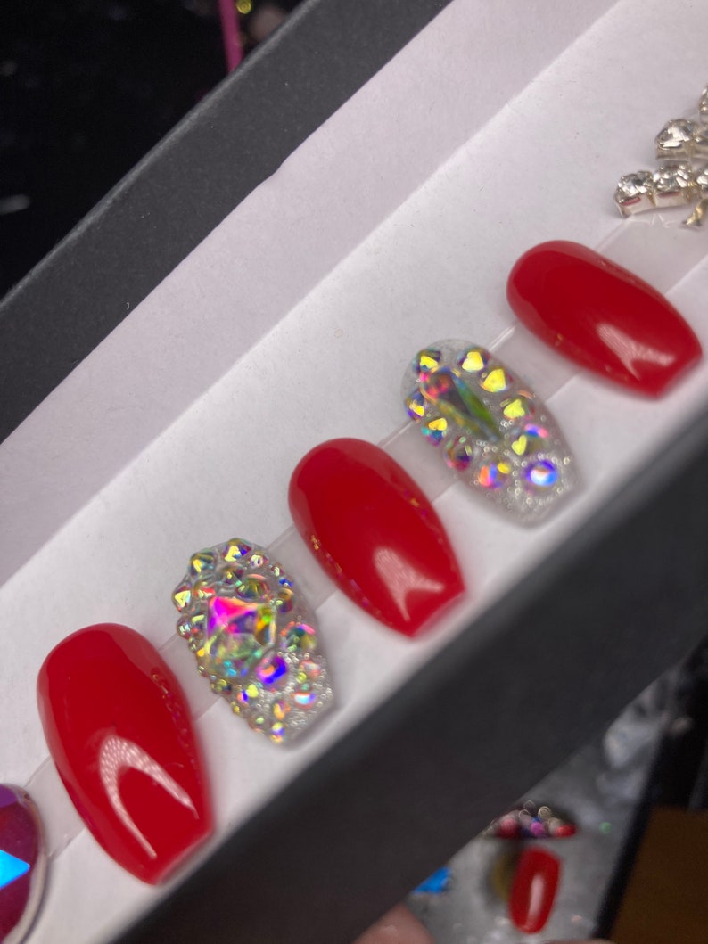 May include: A set of red and rhinestone-encrusted press-on nails. The nails are displayed in a black box with a white interior. The red nails have a glossy finish, while the rhinestone nails sparkle with iridescent gems.