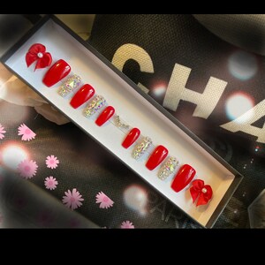 May include: A set of red press-on nails in a black box, with some nails adorned with iridescent rhinestones. Two red bows with pearls are placed at each end of the box. The nails are rectangular shaped.