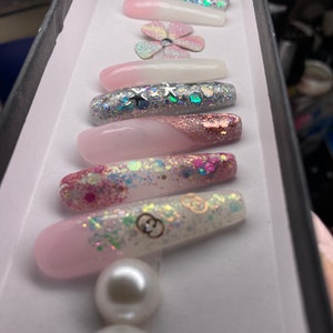May include: A set of long, pink and white press-on nails with glitter and pearl accents. The nails feature a variety of designs, including stars, flowers, and a Gucci logo.