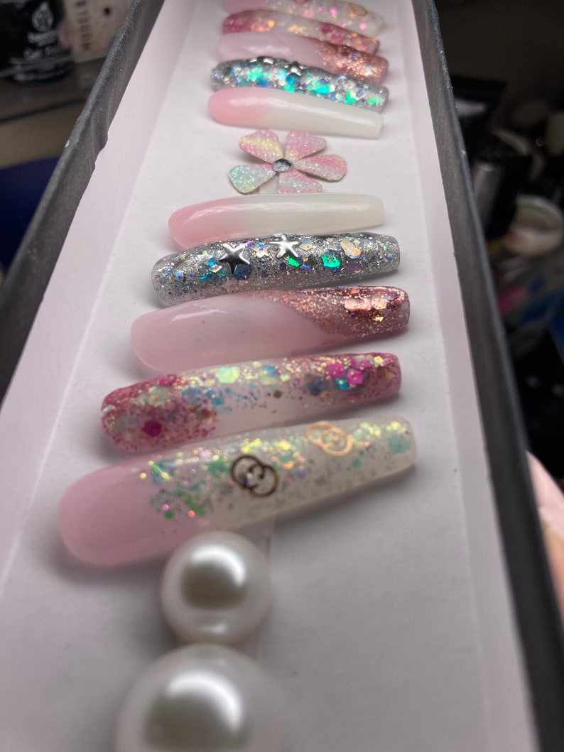 May include: A set of long, pink and white press-on nails with glitter and pearl accents. The nails feature a variety of designs, including stars, flowers, and a Gucci logo.