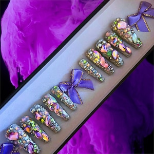May include: A set of ten silver press-on nails with iridescent rhinestones and a lavender bow. The nails are displayed in a white box.