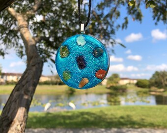 May include: A circular blue pendant necklace with embedded colourful stones. The pendant hangs from a black cord and has a silver-coloured bail. The stones are various colours, including green, brown, and purple. The background is a blurred outdoor scene.