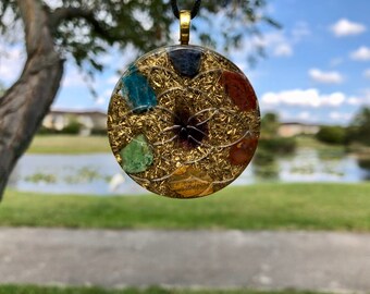 May include: A round pendant with a gold-colored metal frame, featuring colorful gemstones and a central design. The pendant hangs from a black cord with a gold-colored clasp. The background is a blurred outdoor scene with trees and water.