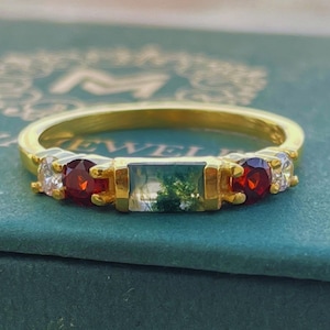 May include: A gold ring with a green stone, two red stones, and two white stones. The ring is on a green box with the word "JEWEL" visible.
