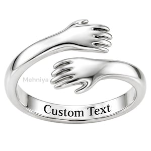 May include: A polished silver-toned open ring featuring two hands reaching towards each other in a hugging design. The band includes the text "Custom Text" engraved on the front surface and the word "Mehniya" printed in small letters near the side.