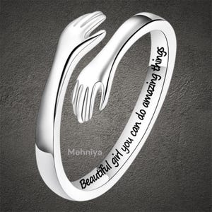 May include: A silver-colored ring with a hugging hands design. The ring features the engraved text "Beautiful girl you can do amazing things". The ring is shown on a gray background.