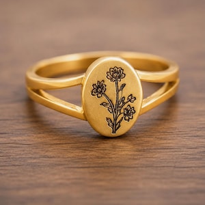 May include: A gold-toned ring featuring an oval-shaped face with a detailed black engraved floral design. The band of the ring is split into two thin sections that connect to the central oval plaque, creating a polished and structured appearance.