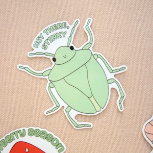 May include: A light green sticker featuring a cartoon stink bug with the text "Hey there, Stinky." The bug has a rounded body, six legs, and two antennae. The sticker is on a light tan background.