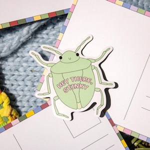 May include: A light green sticker in the shape of a bug with the text "Hey There, Stinky" in pink. The sticker is on a white postcard with a colorful border, resting on a blue knitted surface.