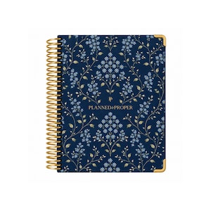 May include: A navy blue spiral-bound planner featuring a repeating pattern of light blue flowers and gold vines. The cover displays the text "PLANNED & PROPER" in gold lettering with a small logo above it. The planner has gold metal corner protectors.