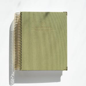 May include: A sage green spiral-bound planner with gold-toned wire binding. The front cover features the embossed text "PLANNED PROPER" with a circular logo above it. The planner includes decorative gold-toned metal corner protectors.