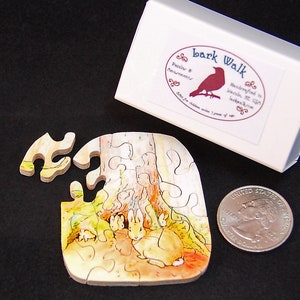 以下が含まれることがあります： 木製の小さなジグソーパズル。木の下にウサギの家族がいます。パズルは「Bark Walk」と書かれた白い箱に入っています。箱の隣にはアメリカの25セント硬貨があります。