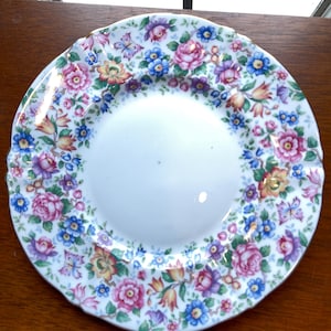 Puede incluir: Un plato de cerámica blanco con un diseño floral. El plato presenta un área central blanca rodeada por un borde de flores coloridas en tonos de rosa, azul, amarillo y morado. El plato tiene un borde festoneado.