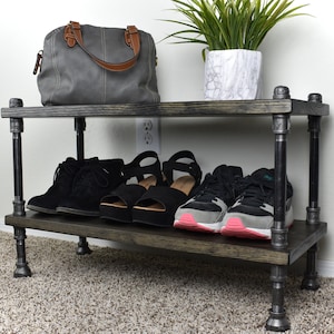 May include: A two-tier storage shelf constructed with dark stained wooden planks and industrial-style dark metal pipe framing. The unit features four vertical pipe supports and holds a gray handbag, a marble-patterned planter, and three pairs of shoes.
