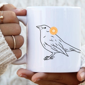 Könnte beinhalten: Eine weiße Keramiktasse mit einer schwarzen Linienzeichnung eines Vogels. Der Vogel blickt nach rechts und hat einen kleinen Kreis mit einem Pfotenabdruck auf seiner Brust. Die Tasse wird von den Händen einer Person gehalten.