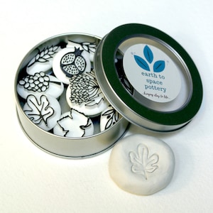 May include: A silver tin with a clear lid containing a set of 10 white clay stamps with black leaf and flower designs. The lid has a label that reads "earth to space pottery bringing clay to life..." A single white clay stamp with a leaf design is sitting next to the tin.