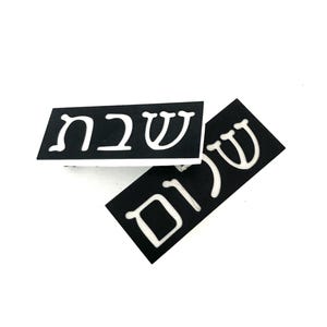May include: Two rectangular black plaques with white Hebrew lettering. The top plaque features the word Shabbat, while the bottom plaque, positioned at an angle, displays the word Shalom. Both items have a clean, high-contrast design.