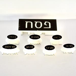 May include: A white and black Passover Seder plate set. The set includes a black sign with white Hebrew text and seven white, oval-shaped dishes with black labels. The labels have Hebrew text.