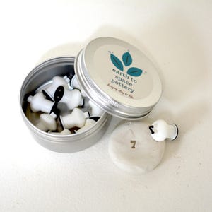May include: A white metal tin with a lid containing small white and black pottery tools. The lid has a sticker with the text "earth to space pottery" and a green leaf design. A small piece of clay with the number "7" imprinted on it is next to the tin.