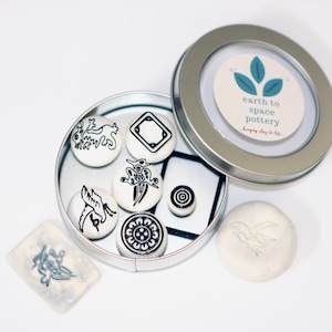 May include: A set of white ceramic magnets with black graphic designs, displayed in an open silver tin. The tin includes a label with the text "earth to space pottery". A small, rectangular tile with a matching design is also present.