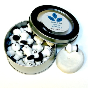May include: A round metal tin with a clear lid containing white ceramic stamps with black accents. The lid has a label that reads "earth to space pottery...bringing clay to life..."  A white ceramic stamp is sitting on a small piece of white clay.