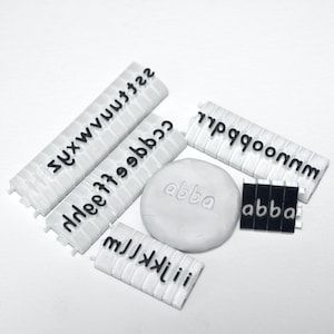 May include: White plastic tiles with black letters spelling out the alphabet. The tiles are arranged in rows and columns. There is a small white clay block with the word "abba" imprinted on it.