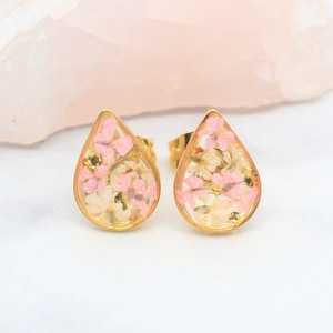 May include: A pair of gold-colored teardrop stud earrings. Each earring features a clear resin interior filled with small pink and white flowers and gold flakes. The earrings are set against a soft pink background.