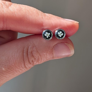 Könnte beinhalten: Ein Paar runde Ohrstecker. Jeder Ohrring hat einen schwarzen Hintergrund mit einem weißen Blumendesign und einem silbernen Rand. Die Ohrringe werden zwischen den Fingern gehalten.