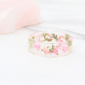 May include: A clear resin ring with pink and green pressed flowers embedded inside.