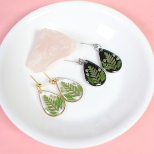 Puede incluir: Dos pares de pendientes en forma de lágrima con helechos y flores prensados en su interior. Un par tiene resina transparente y detalles dorados, el otro tiene resina negra y detalles plateados.