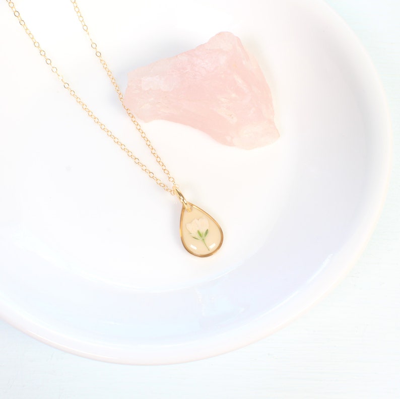 May include: A gold chain necklace with a teardrop-shaped pendant featuring a delicate white flower pressed inside clear resin.