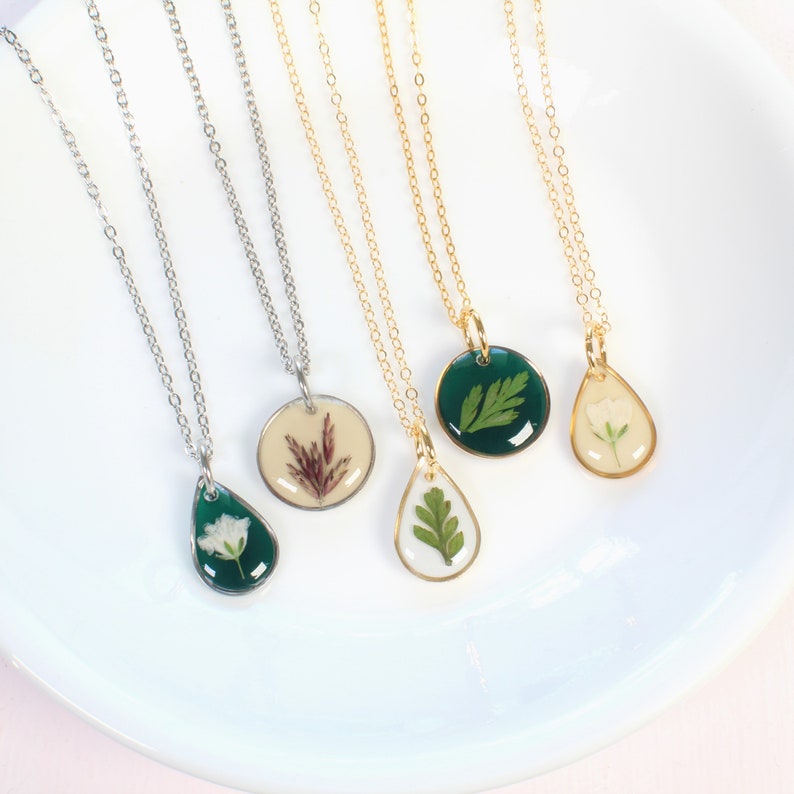 May include: Four necklaces with different pendants featuring pressed flowers and leaves encased in resin. The pendants are in the shapes of a circle and a teardrop. The chains are silver and gold.