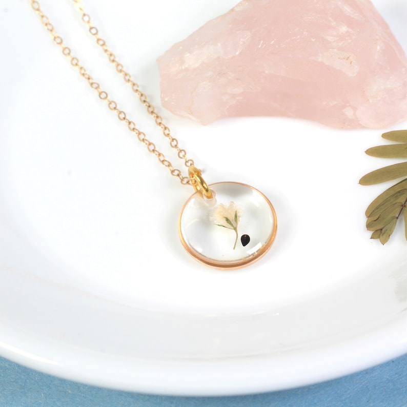 May include: A gold chain necklace with a round pendant featuring a small white flower pressed inside clear resin.