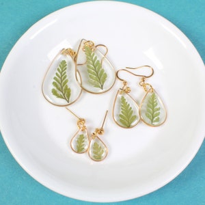 May include: A set of four gold teardrop earrings with pressed green ferns inside the resin. The earrings are on a white plate.
