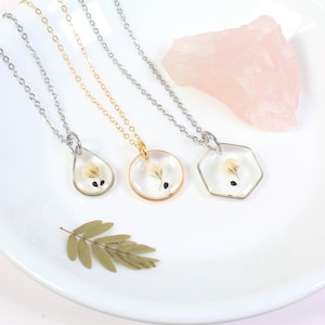 May include: Three delicate necklaces with different pendant shapes: a teardrop, a circle, and a hexagon. Each pendant features a pressed flower encased in clear resin. The necklaces are on a white surface with a pink rock in the background.