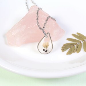 May include: A silver chain necklace with a teardrop-shaped pendant containing a pressed flower and seeds. The pendant is clear and the flower is white.